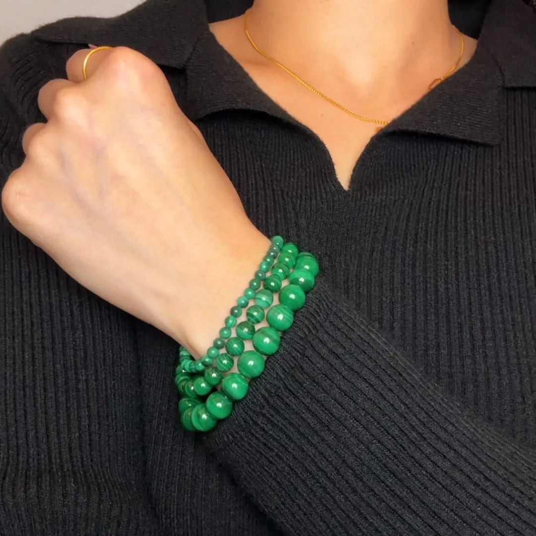 Trois bracelets en perles de malachite verte de différentes tailles, portés sur le poignet d'une femme vêtue d'un pull noir.