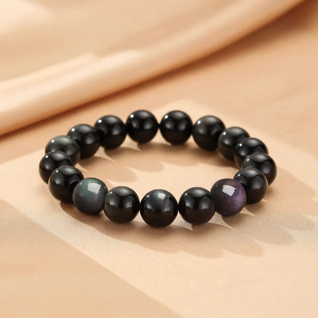 Bracelet fait de perles d'obsidienne arc-en-ciel noires et irisées, disposées en cercle sur un fond texturé beige.