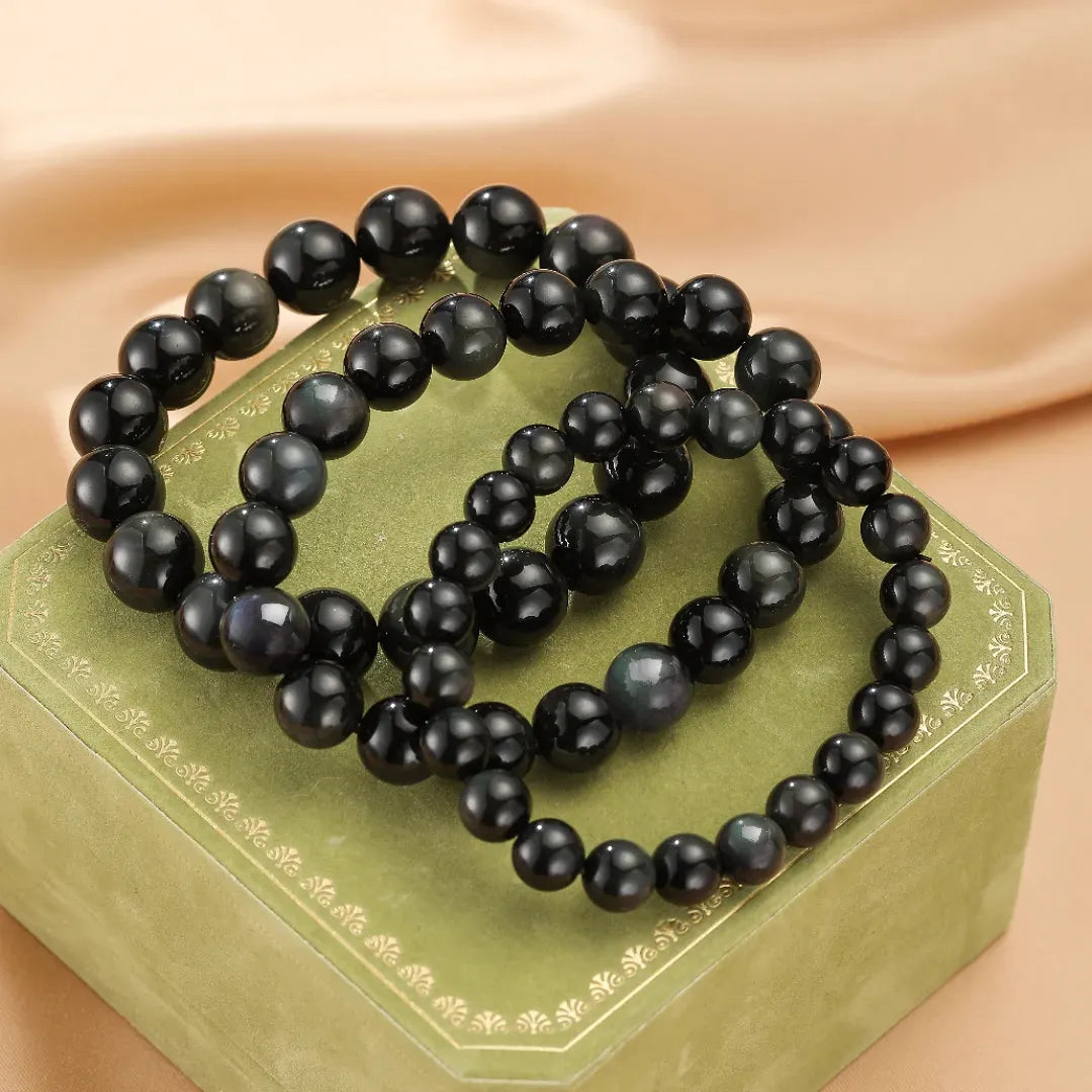 Trois bracelets en perles d'obsidienne arc-en-ciel "Profondeur Mystérieuse" reposant sur une boîte verte ornée.