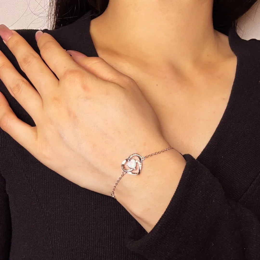 Bracelet en argent avec un pendentif en forme de cœur double orné d'une opale blanche et de petites pierres scintillantes, porté au poignet d'une femme.