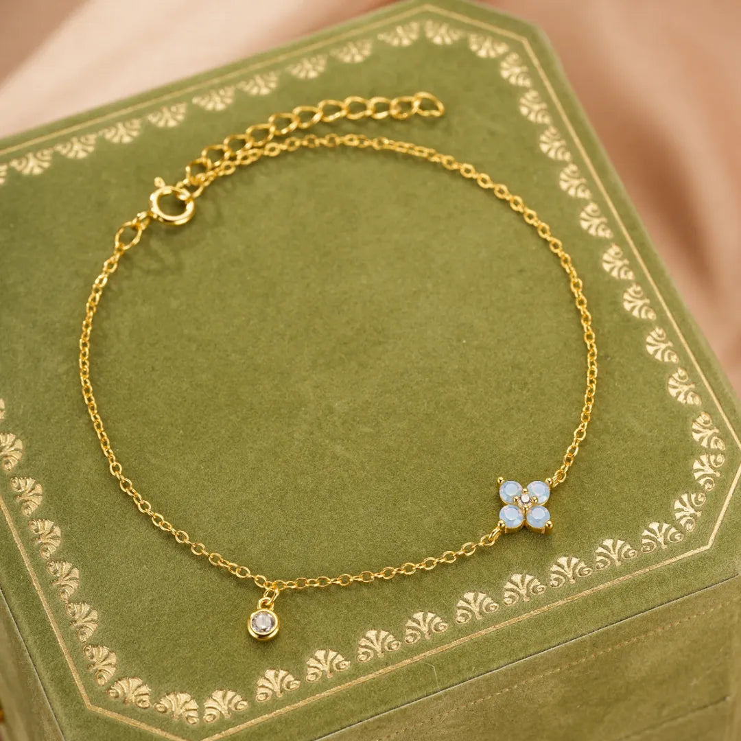 Bracelet fin en argent doré à l'or, orné d'une petite opale en forme de fleur de quatre pétales et d'un cristal brillant, présenté sur un écrin vert.
