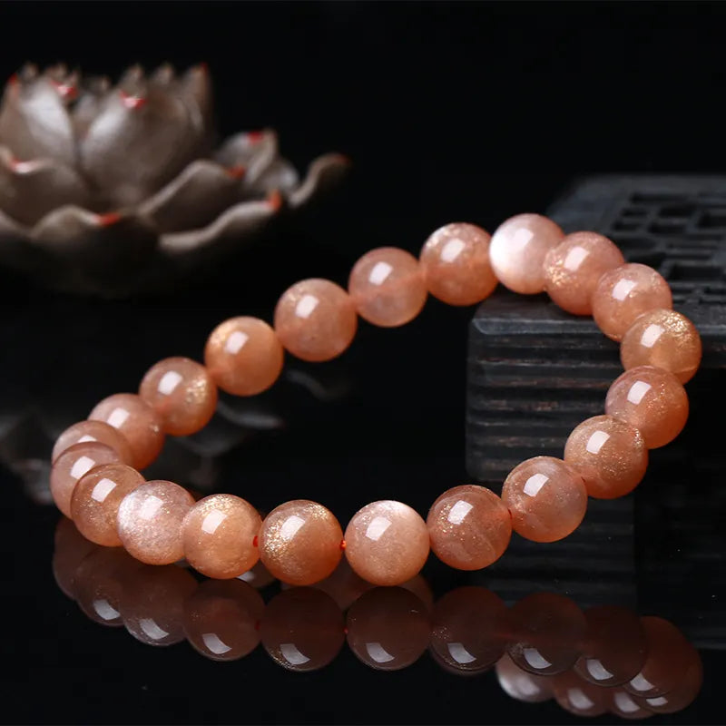Bracelet en pierres de soleil "Élégance Solaire" aux reflets dorés chatoyants, monté sur un fil rouge discret. Gros plan sur le bijou posé sur un support en bois sombre, avec un objet décoratif flouté en arrière-plan.
