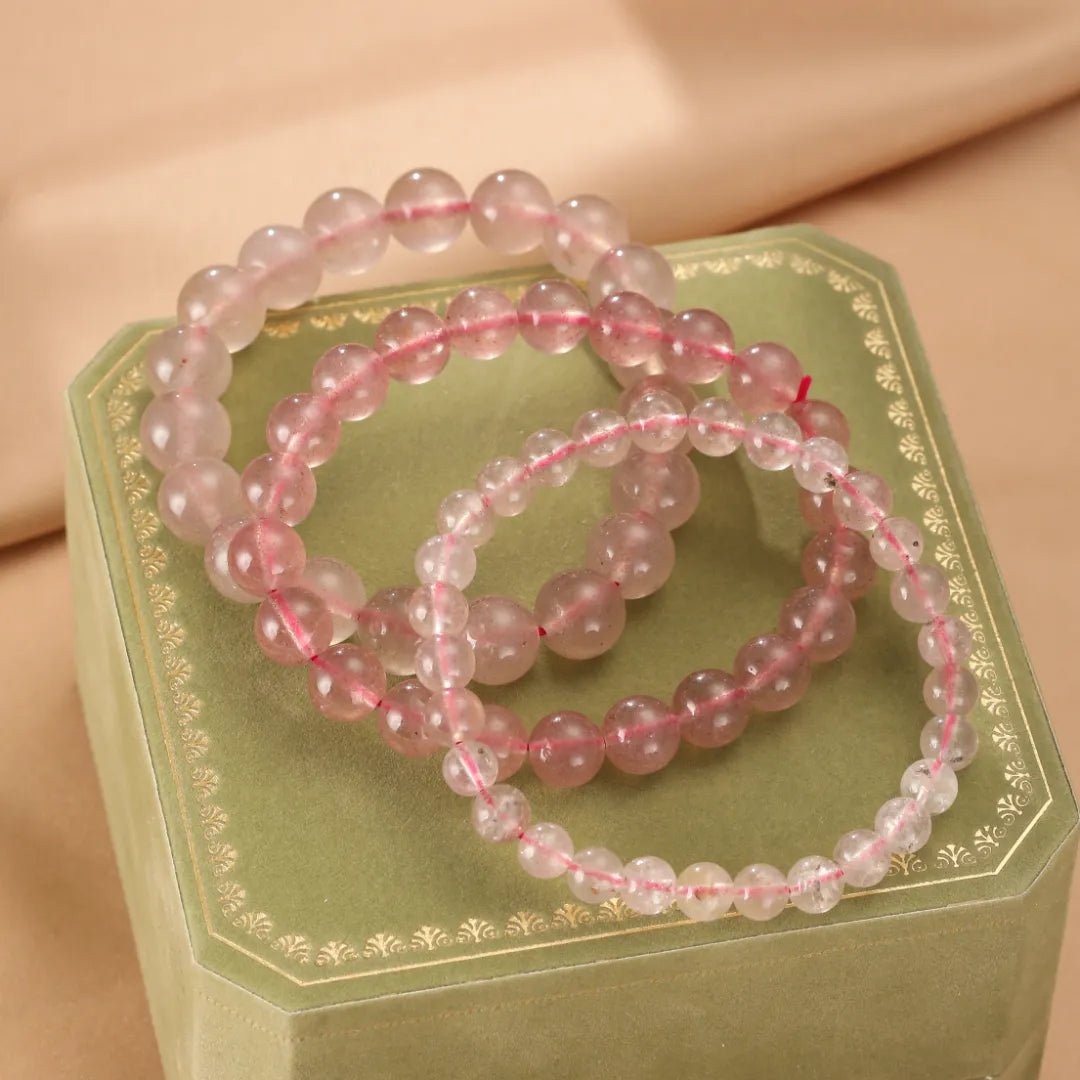 Trois bracelets en quartz fraise aux tons roses doux, présentés sur une boîte verte ornée de dorures.
