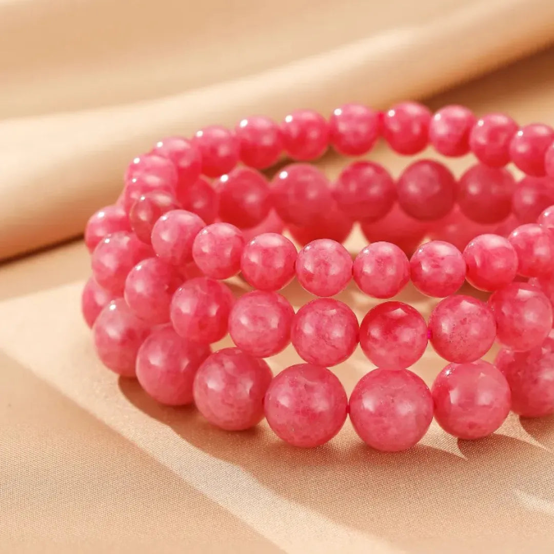 Trois bracelets faits de perles rondes de rhodonite rose tendre, disposés en couches sur un fond beige texturé.