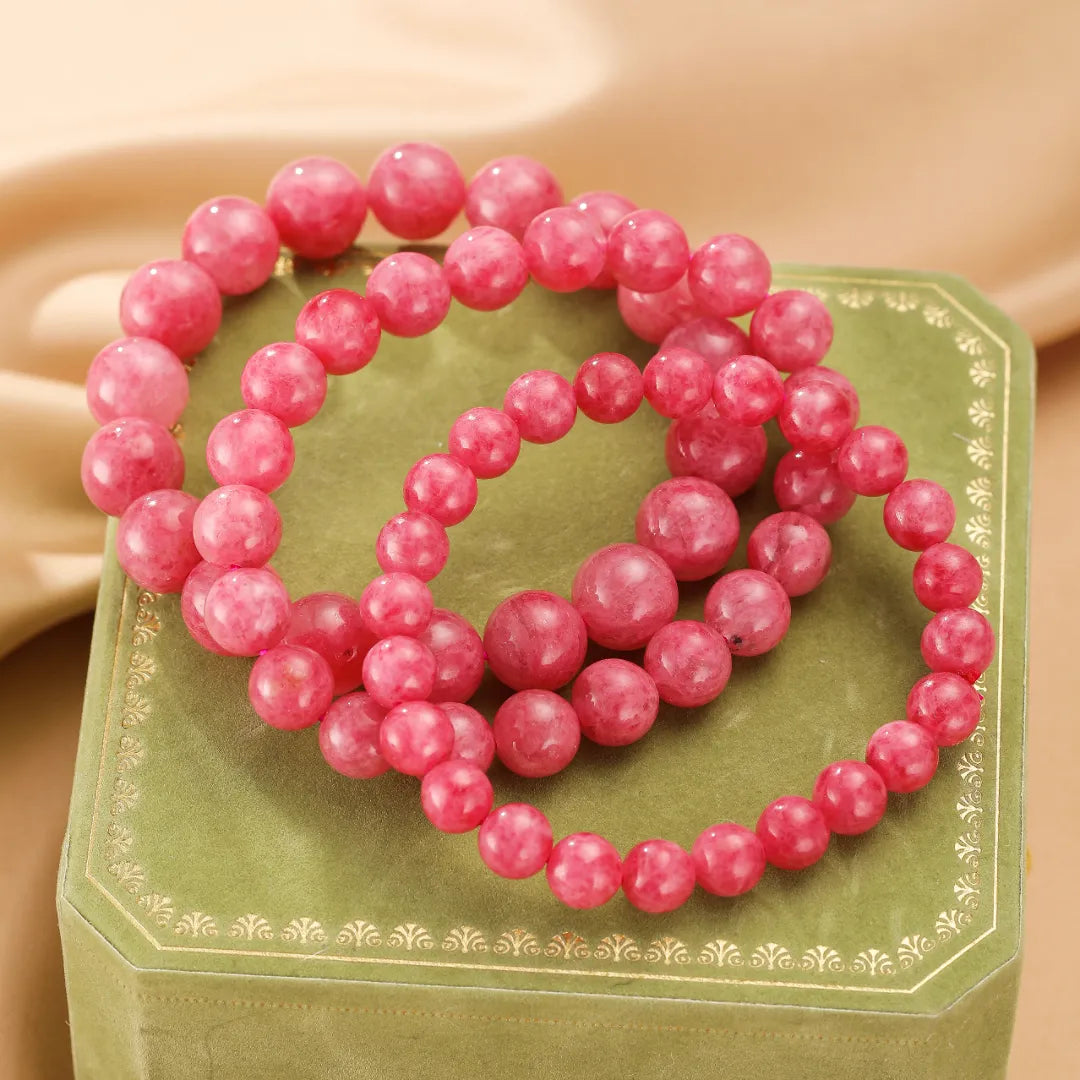 Trois bracelets en perles de rhodonite rose pâle disposés en cercle sur une boîte vert émeraude ornée de motifs dorés.