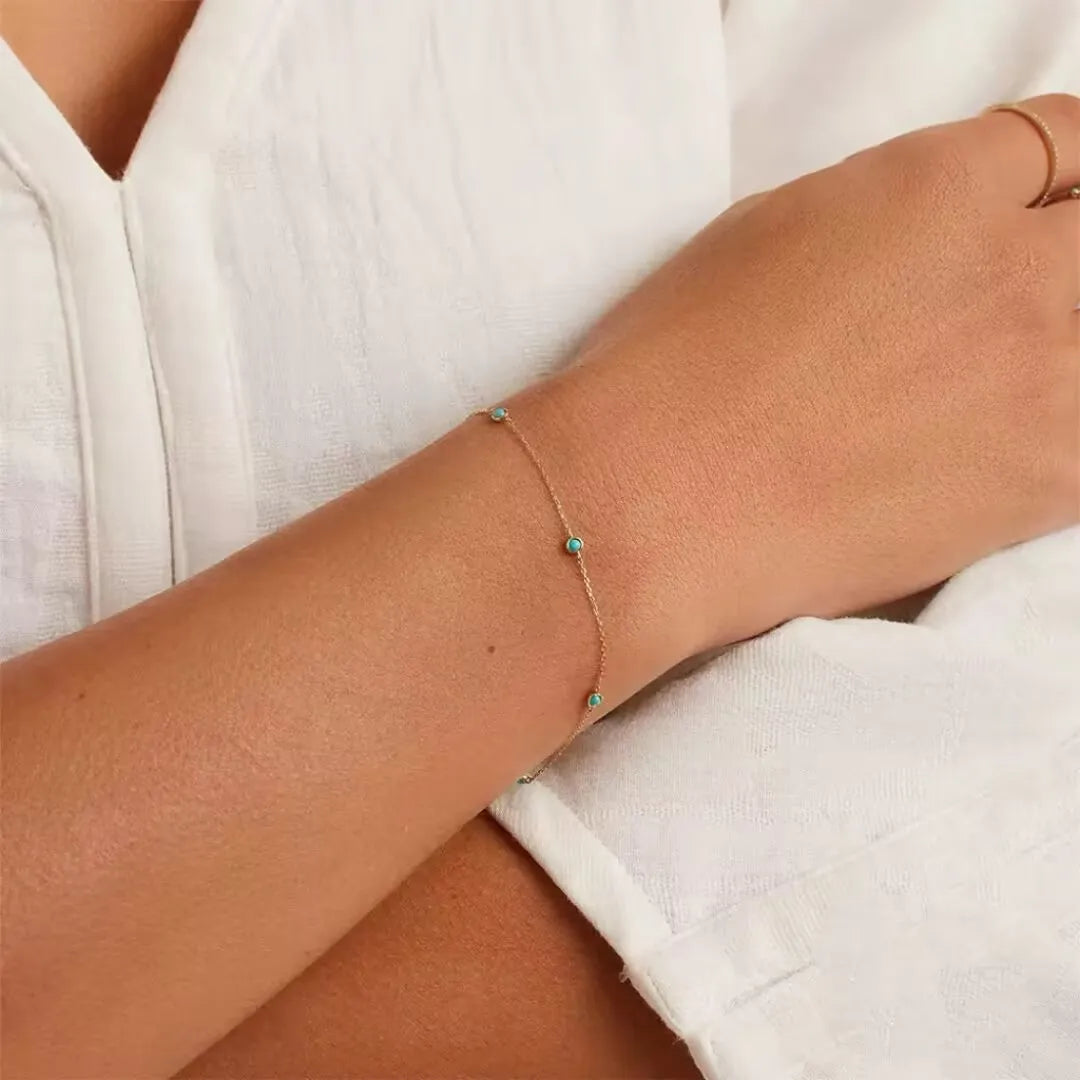 Bracelet fin en argent doré or avec plusieurs petites pierres turquoise disposées le long de la chaîne, porté au poignet d'une femme.