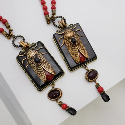 Deux colliers "Beauté Naturelle" avec pendentifs rectangulaires noirs ornés d'une cigale dorée, d'une pierre rouge et de perles rouges et noires.