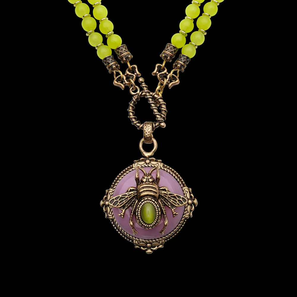 Collier fantaisie avec un pendentif en forme d'abeille dorée sur fond rose, orné d'une petite pierre verte, et deux rangées de perles jaunes.