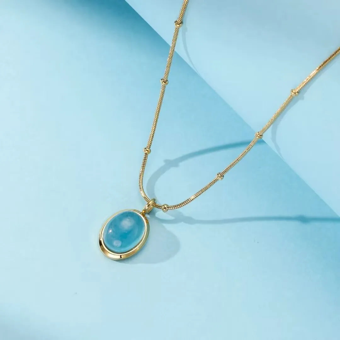 Collier plaqué or avec un pendentif ovale en aigue-marine bleu clair sur une chaîne délicate ornée de petites perles.