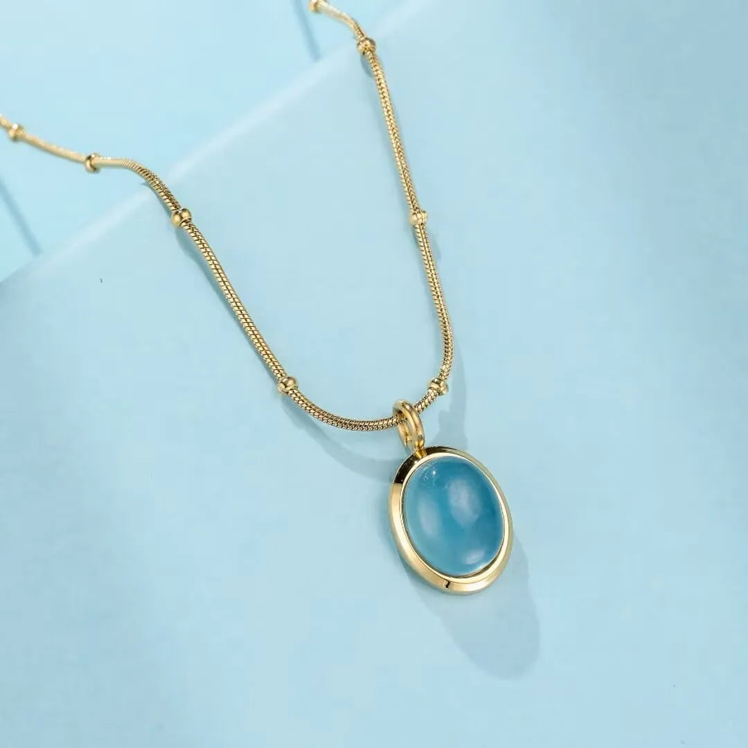 Collier en acier titane doré à l'or 18 carats avec un pendentif ovale en aigue-marine bleue claire, sur fond bleu clair.