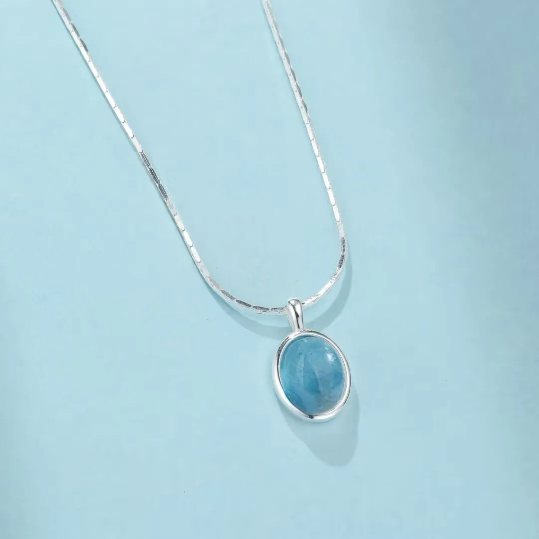 Collier en argent 925 avec un pendentif ovale en aigue-marine bleu clair, sur fond bleu pastel.