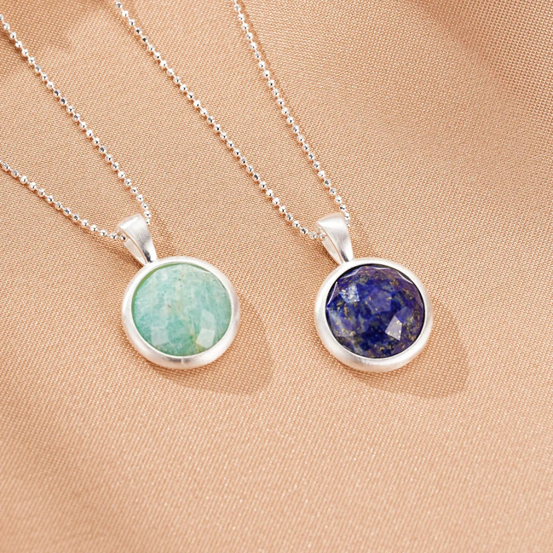 Deux colliers "Profondeur Céleste" en argent, l'un avec une pierre d'amazonite vert pâle, l'autre avec une pierre de lapis-lazuli bleu profond, sur un fond beige texturé.