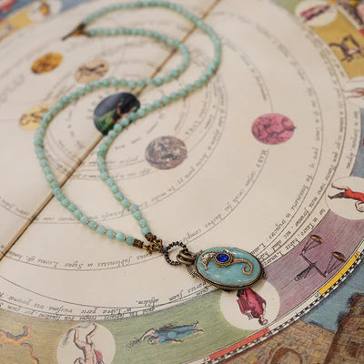 Collier "Voyage Marin" en amazonite avec un pendentif ovale bleu clair orné d'un hippocampe bleu incrusté, posé sur un document ancien illustrant un système planétaire.