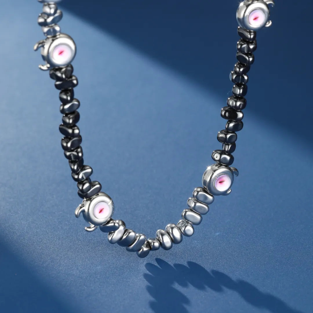 Collier pour homme en hématite noire avec des pendentifs "œil" argentés au centre rose et des perles argentées et noires. Le collier "Regard Hypnotique" est présenté sur un fond bleu.