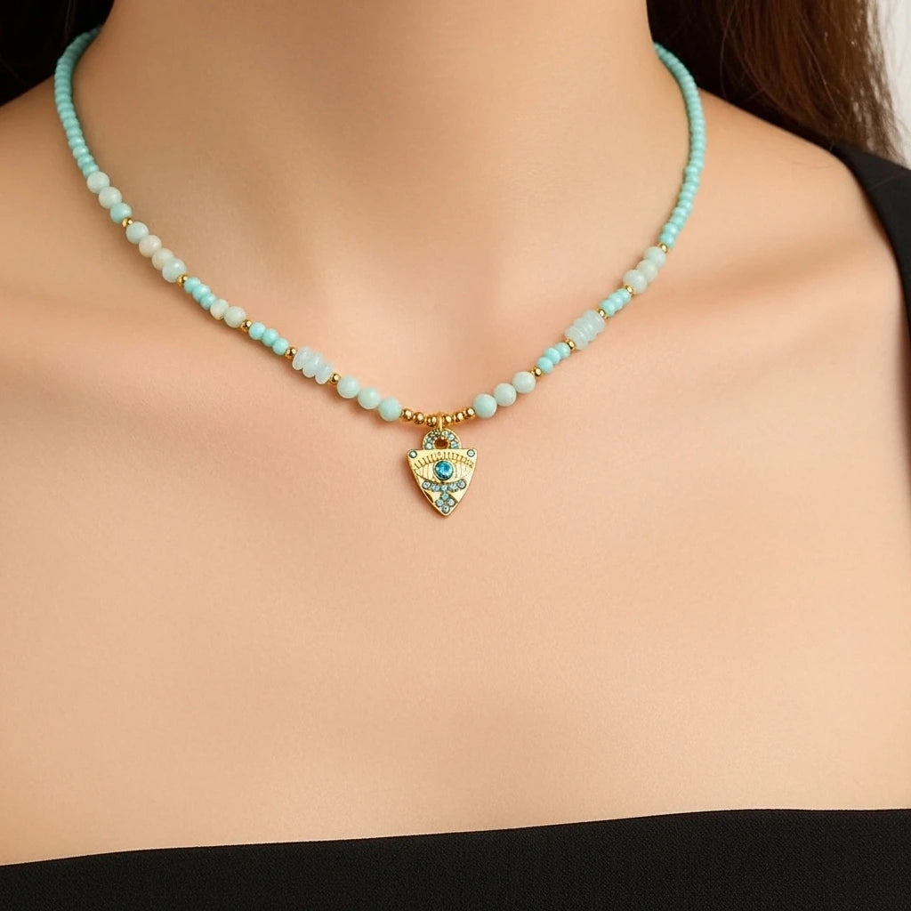 Collier "Œil Apaisant" en amazonite avec un pendentif en forme de bouclier doré orné d'un œil bleu et de petites pierres bleues.