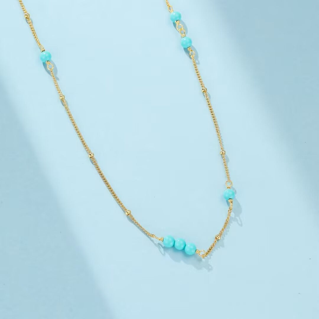 Collier "Dunes de Charme" en argent doré or, orné de perles turquoise et de petites billes dorées, sur fond bleu clair.