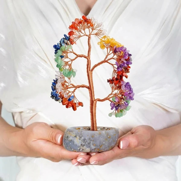 Arbre de vie 7 chakras décoratif avec des pierres colorées dans un pot en pierre grise, tenu dans des mains.