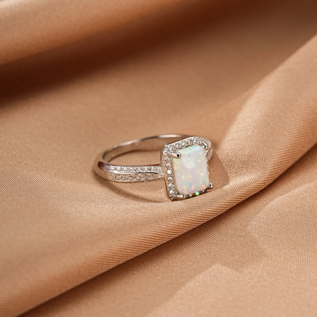 Bague en argent avec une opale rectangulaire blanche entourée de petites pierres scintillantes, posée sur un tissu satiné beige.
