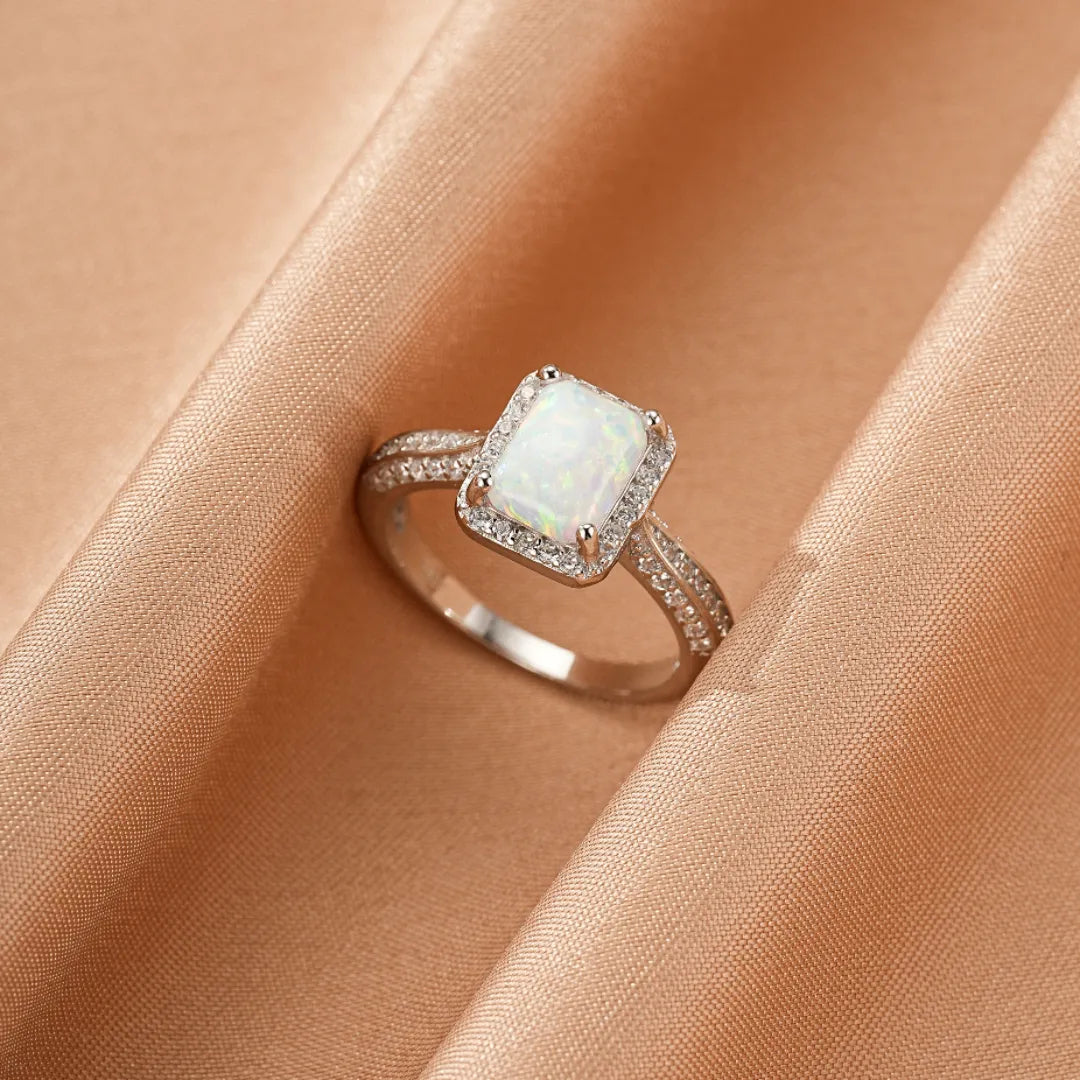 Bague en argent avec une opale rectangulaire entourée de petits cristaux scintillants, posée sur un tissu couleur pêche.