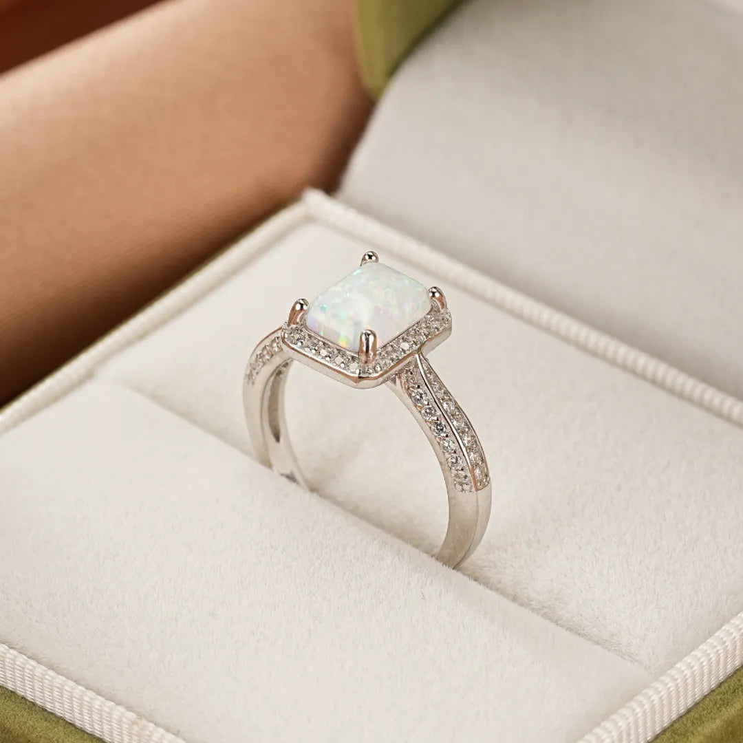 Bague en argent avec une opale centrale de forme rectangulaire entourée de petits cristaux scintillants, posée dans un écrin.