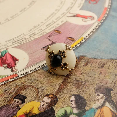 Bague en argent ornée d'une pierre d'agate noire centrale, entourée de motifs floraux dorés, posée sur une illustration ancienne.