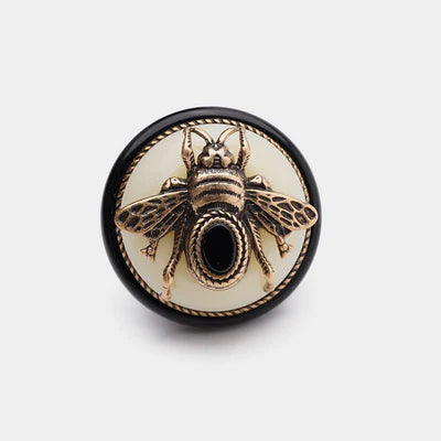 Bague "Reine Noire" avec une abeille en métal doré sur une agate noire, posée sur un fond blanc entouré d'un cordon tressé et d'un anneau noir.