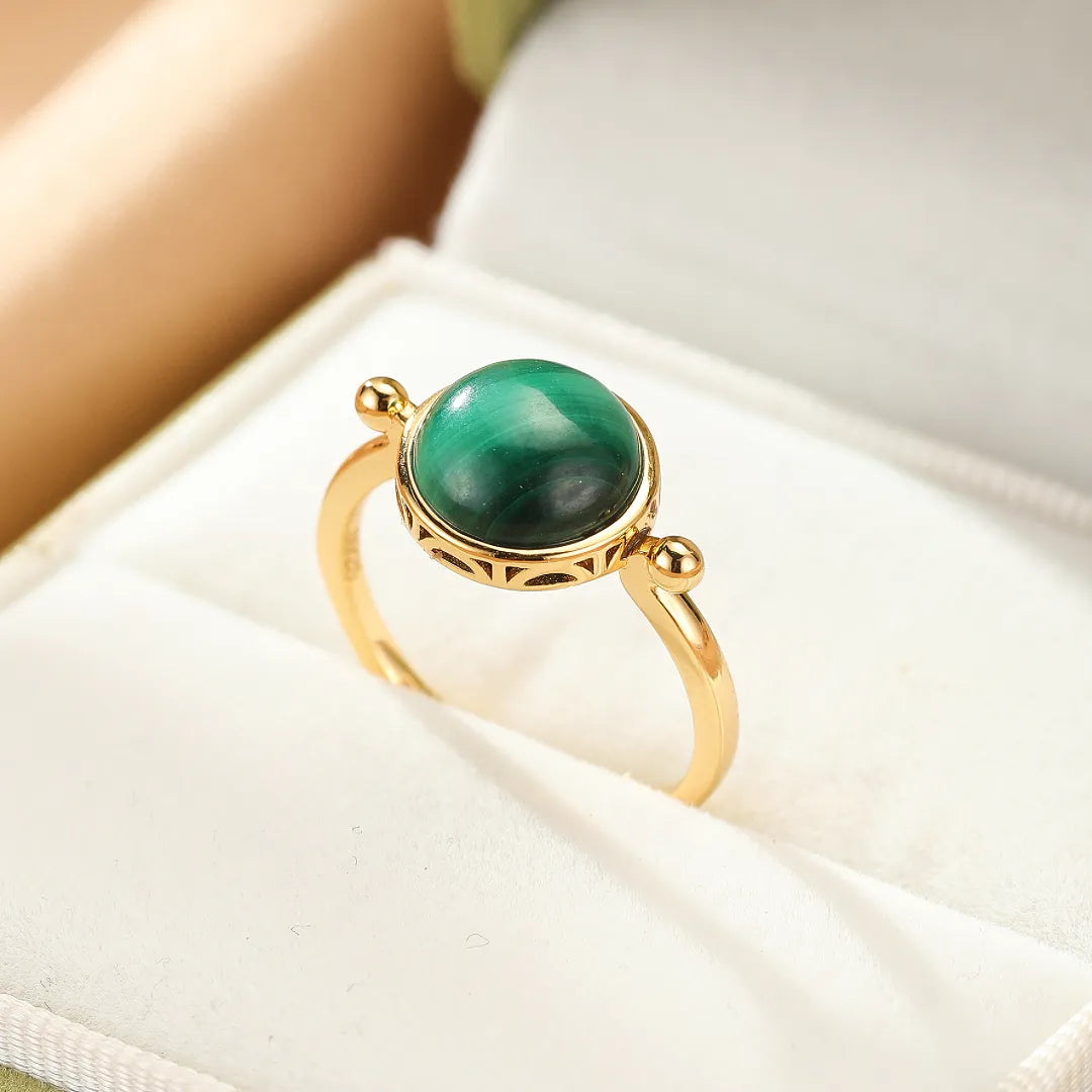 Bague en argent doré avec une pierre de malachite ronde au centre, sertie sur un anneau fin orné de motifs géométriques discrets.