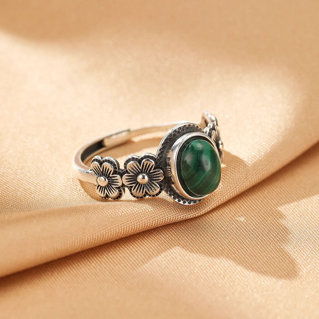 Bague en argent ornée d'une pierre de malachite ovale et de motifs floraux gravés sur le côté, posée sur un tissu beige satiné.