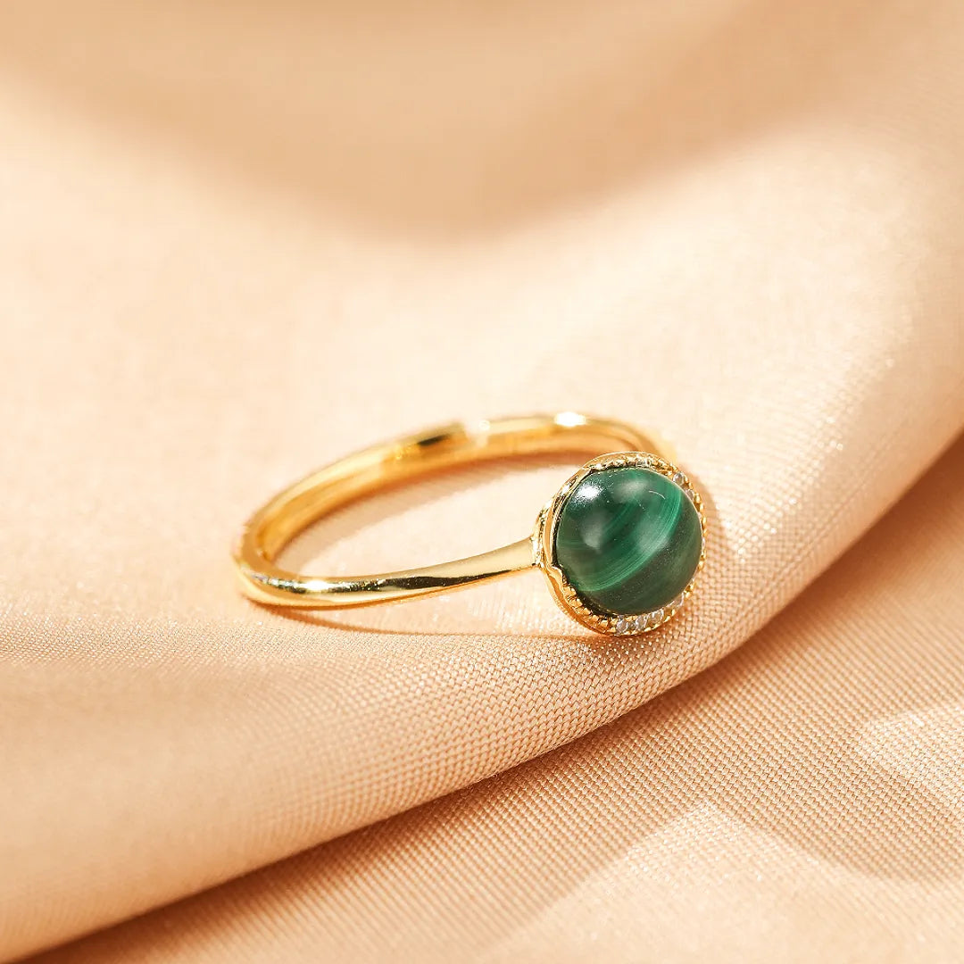 Bague en argent doré sertie d'une pierre de malachite verte ronde et de petites pierres scintillantes, sur un fond en tissu beige satiné.