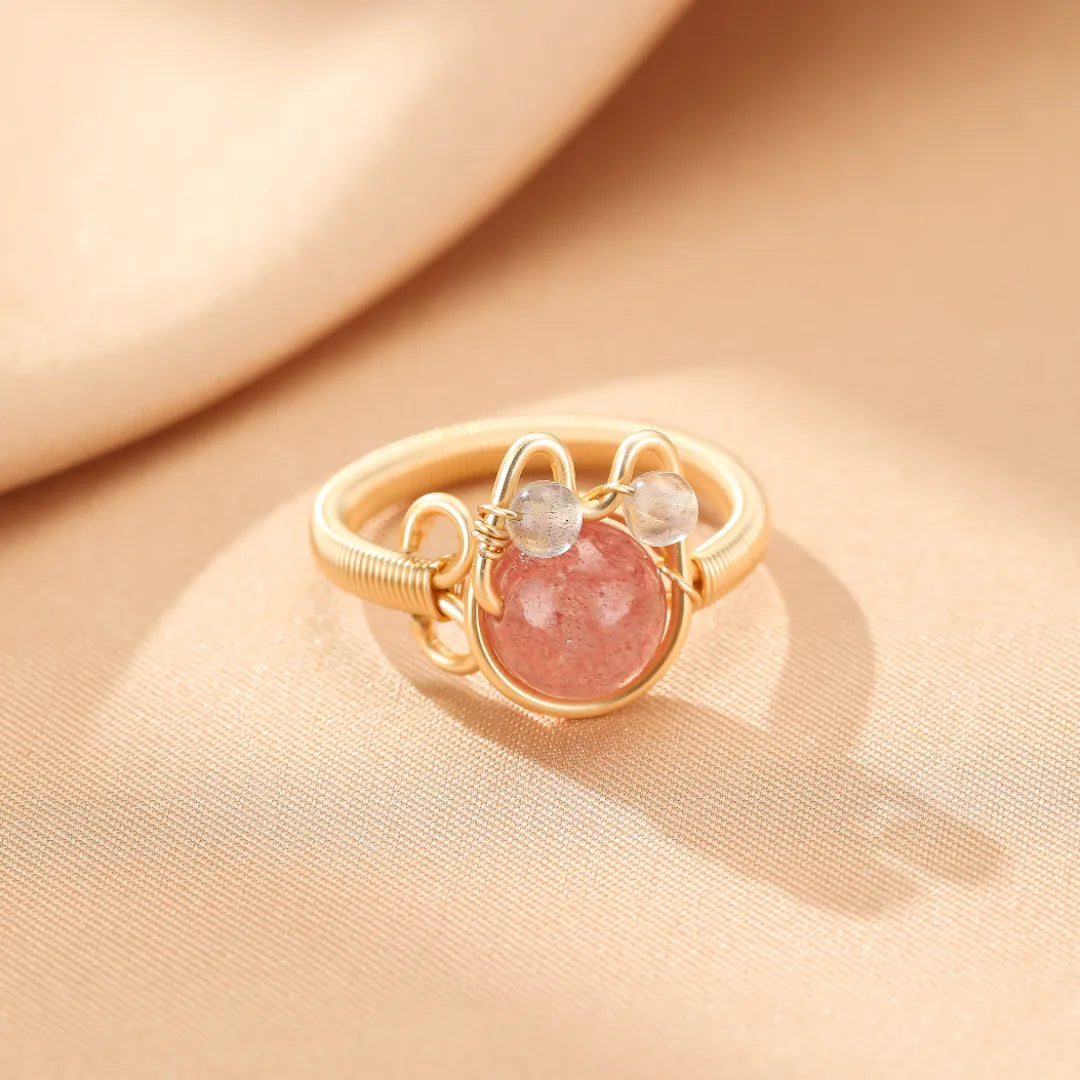 Bague en fil doré orné d'une pierre rose centrale et de deux petites pierres grises, évoquant la forme d'un chaton joyeux.