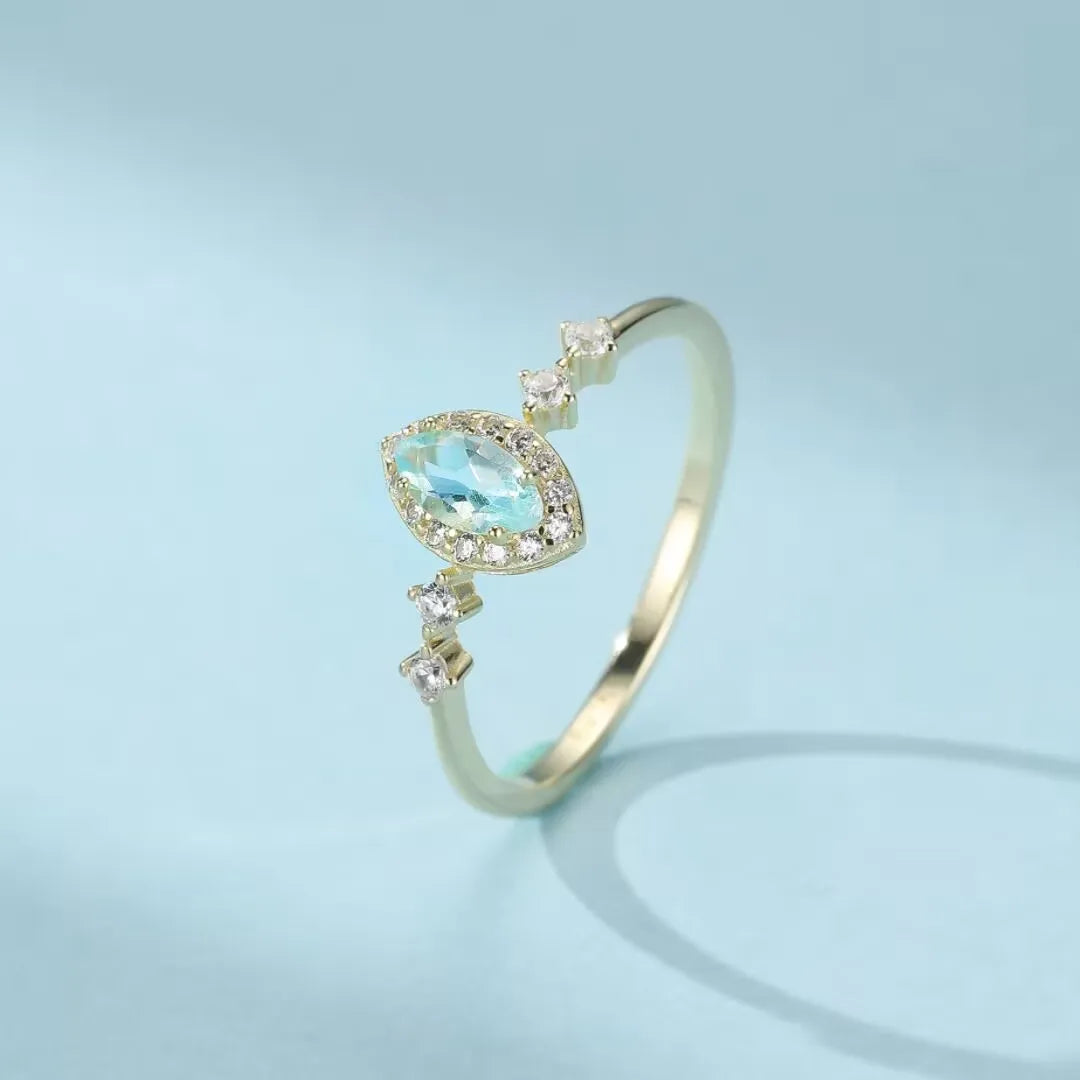 Bague en argent 925 sertie d'une topaze bleue en forme de marquise entourée de petits zirconiums blancs, sur un fond bleu clair.