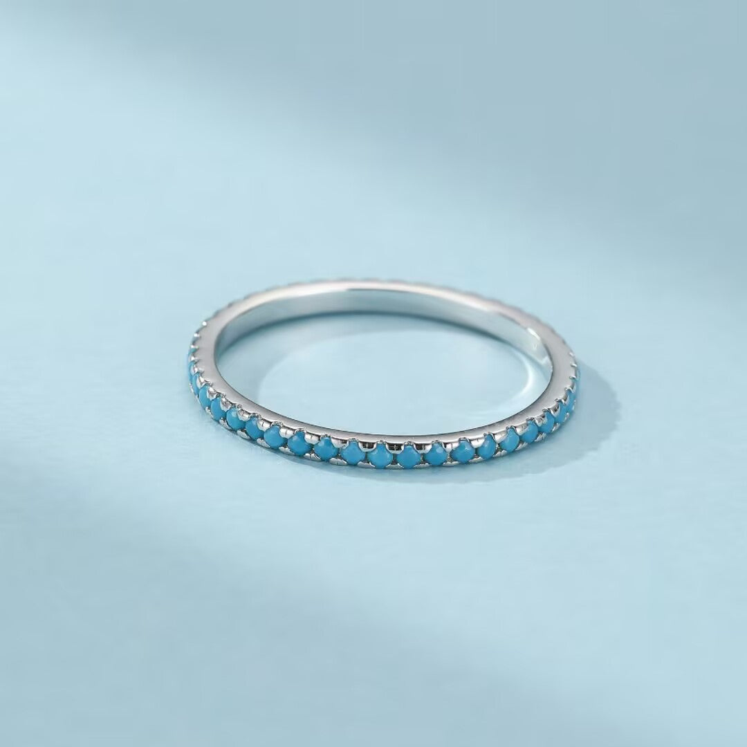 Bague fine en argent 925 sertie de petites pierres de turquoise bleues sur tout son contour, sur fond bleu clair.