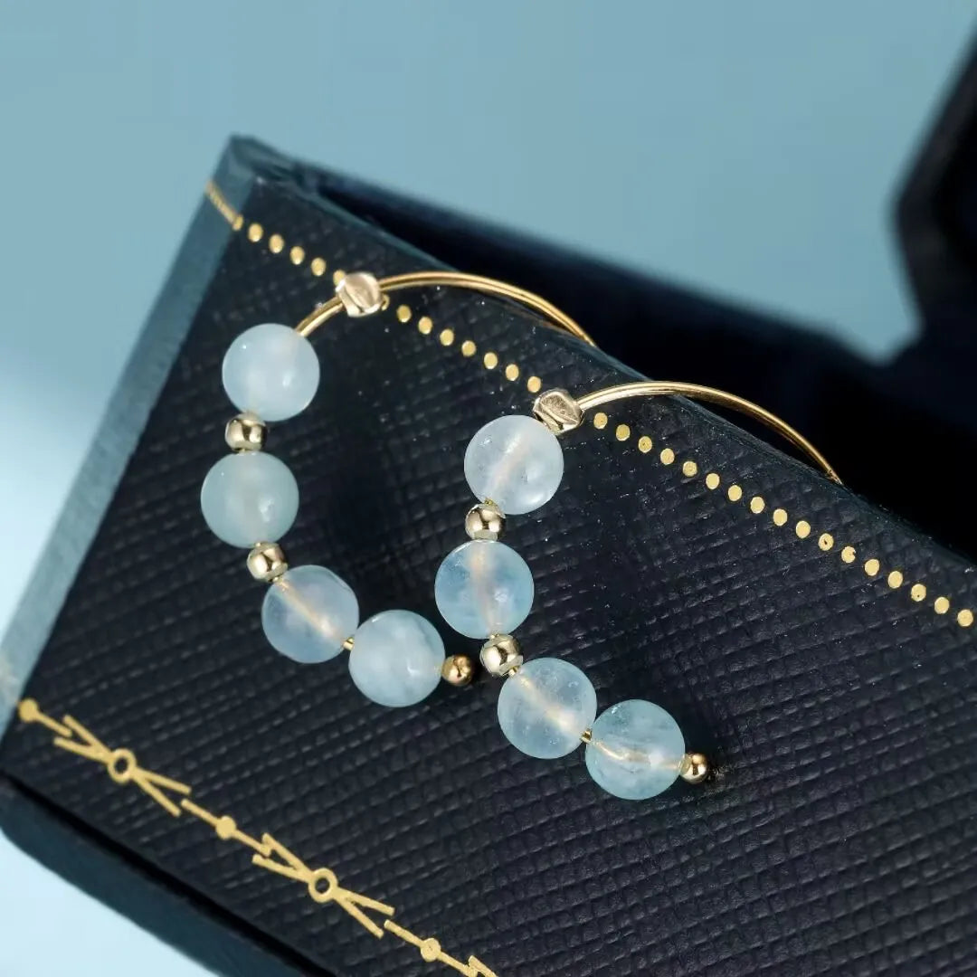 Boucles d'oreilles "Brume Céleste" en aigue-marine et or, présentées sur un écrin noir orné de bijoux noir avec un fond bleu ciel bleu en arrière-plan.