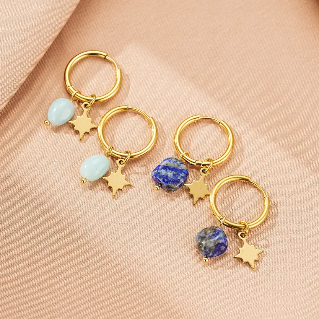 Quatre paires de boucles d'oreilles créoles dorées ornées de breloques étoile et de pierres naturelles bleues (Lapis Lazuli) ou bleu clair (Amazonite), présentées sur un fond rose poudré.