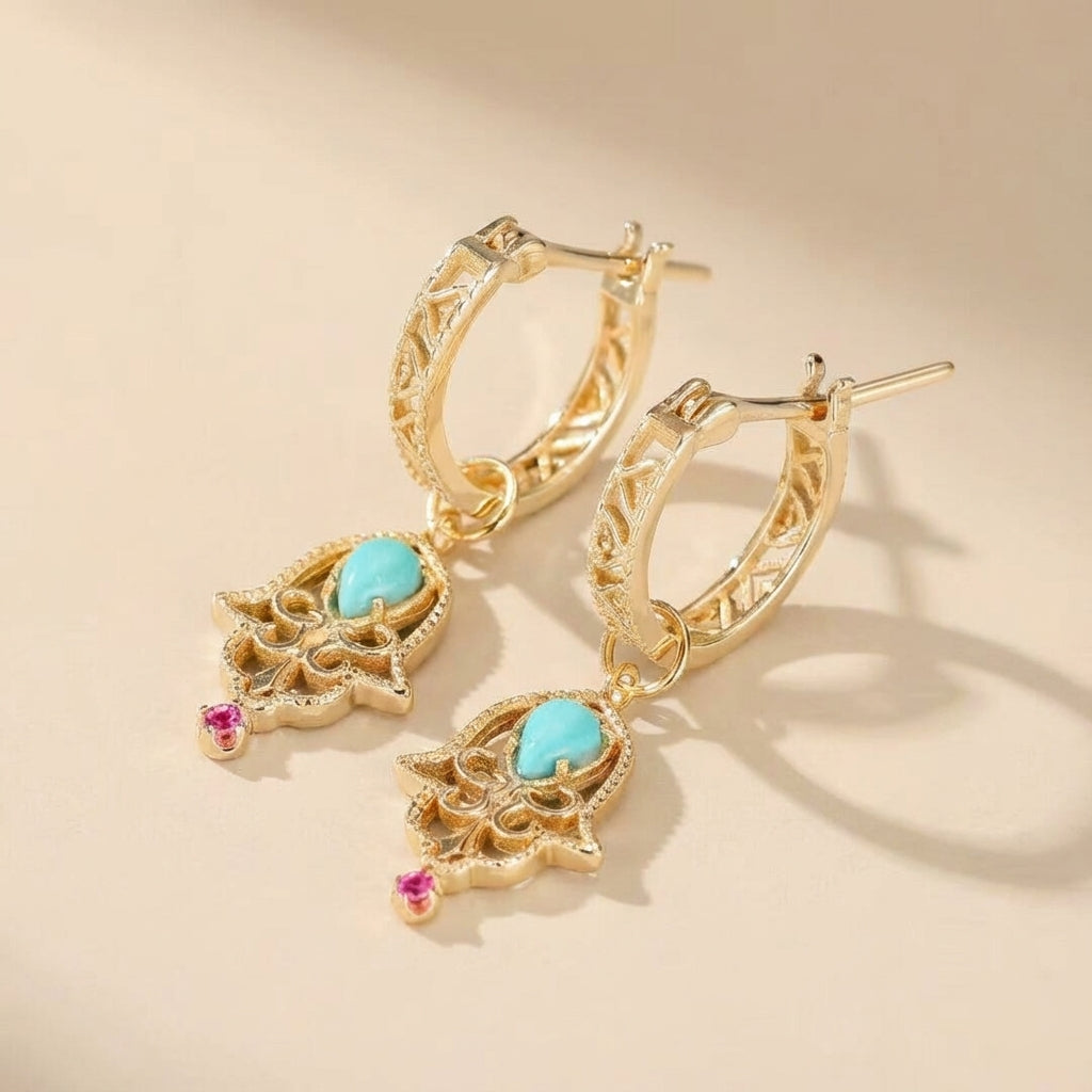 Boucles d’oreilles "Aura Ancestrale" en argent doré, ornées d'une turquoise centrale et de petites pierres roses, avec un design inspiré de la main de Fatma.