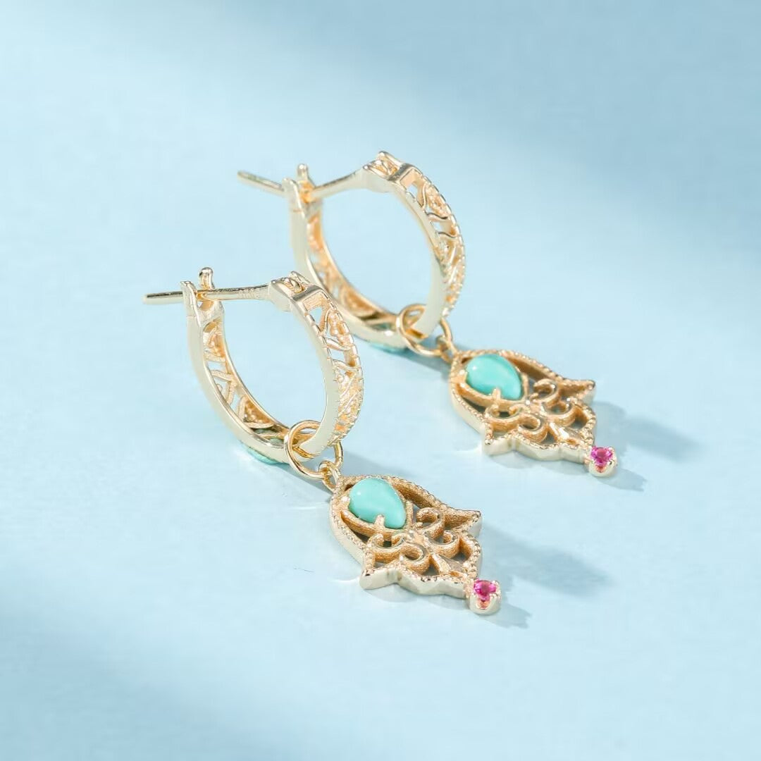 Boucles d’oreilles pendantes en argent doré ornées de turquoise et de petites pierres roses, style main de Fatma, sur fond bleu clair.