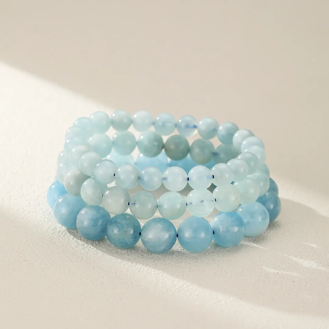 Trois bracelets en perles d'aigue-marine aux teintes bleues claires et douces, empilés les uns sur les autres sur un fond clair.