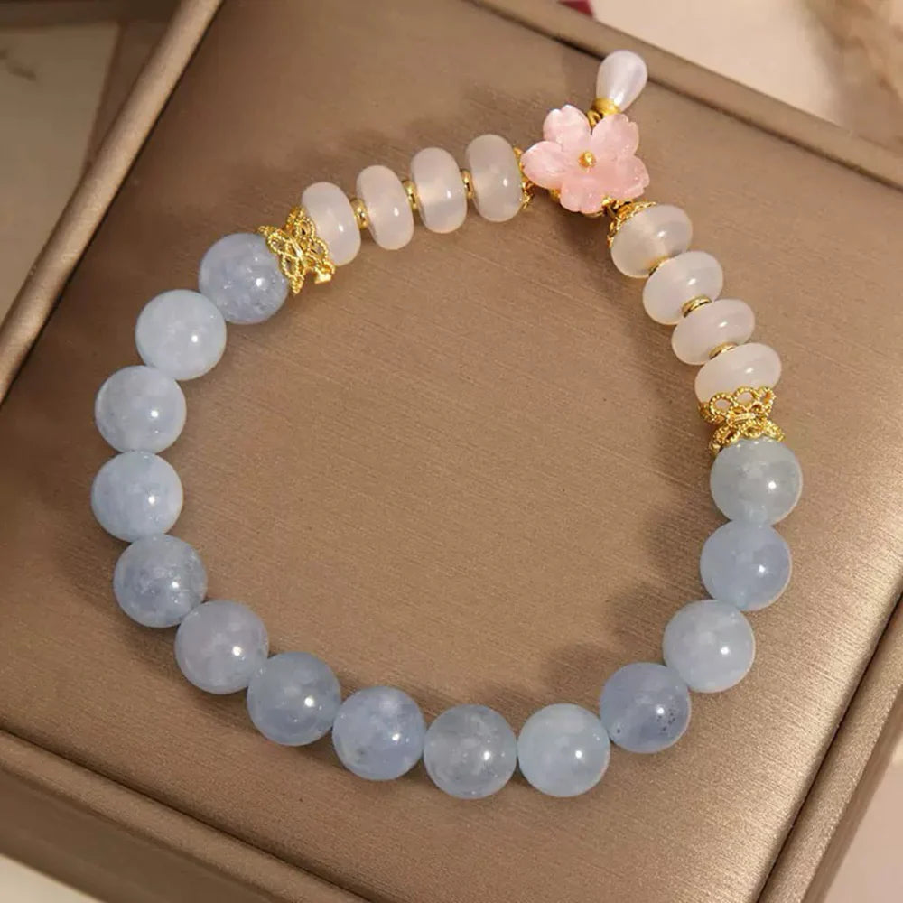 Bracelet "Pureté de Matin" fait de perles d'aigue-marine bleues et de perles d'agate blanches, orné d'une fleur rose et de détails dorés.