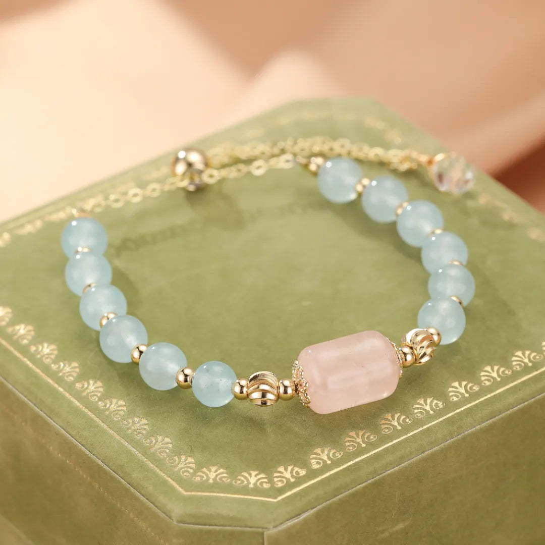 Bracelet "Souffle Doux" fait de perles d'aigue-marine bleu pâle et d'une perle centrale rectangulaire en quartz rose, rehaussé de petites perles dorées, sur un écrin vert.
