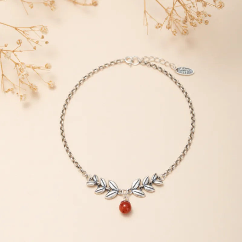 Bracelet de cheville en argent avec une chaîne fine ornée de feuilles stylisées et d'une petite agate rouge suspendue.