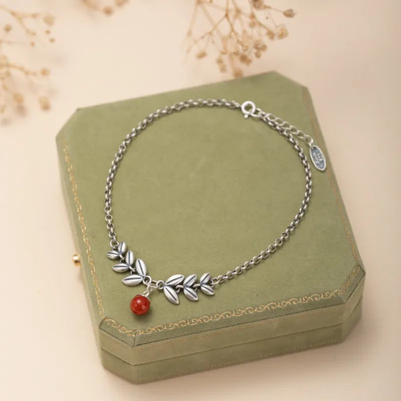 Bracelet de cheville en argent orné de feuilles stylisées et d'une petite agate rouge, reposant sur une boîte à bijoux verte.