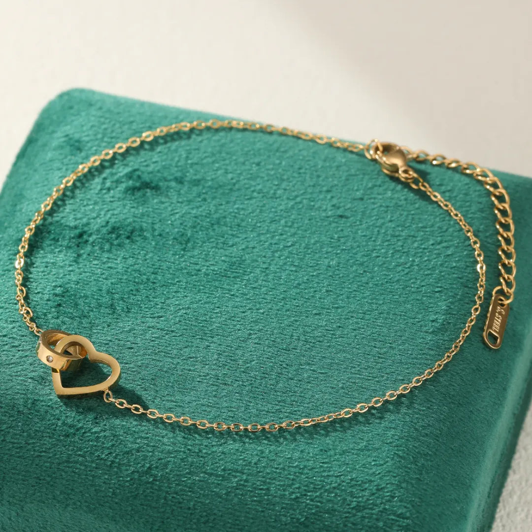 Bracelet de cheville doré avec deux cœurs entrelacés et un petit diamant, sur un fond vert.