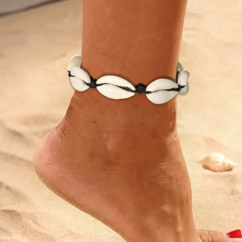 Bracelet de cheville "Douceur Océan" orné de coquillages blancs naturels sur une cheville bronzée, posé sur un sable fin avec une autre coquille en arrière-plan.