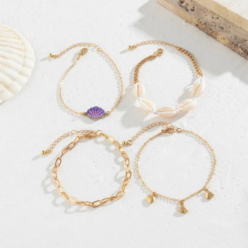 Ensemble de quatre bracelets de cheville dorés, dont un avec un pendentif coquillage violet, un avec plusieurs petits coquillages blancs et deux autres avec des maillons ou des breloques dorées.