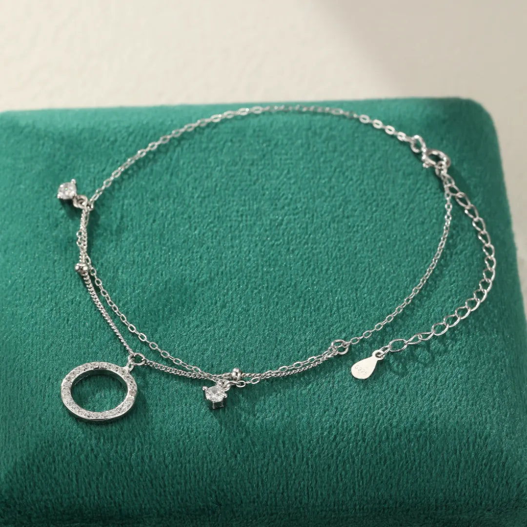 Bracelet de cheville double chaîne argent avec un pendentif rond serti de zircons et deux petites pierres scintillantes.