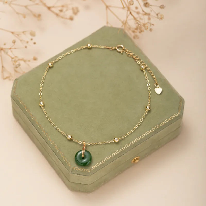 Bracelet de cheville avec un pendentif rond en jade vert foncé, monté sur une chaîne fine en argent doré ornée de petites perles dorées. Le bijou est posé sur une boîte verte.