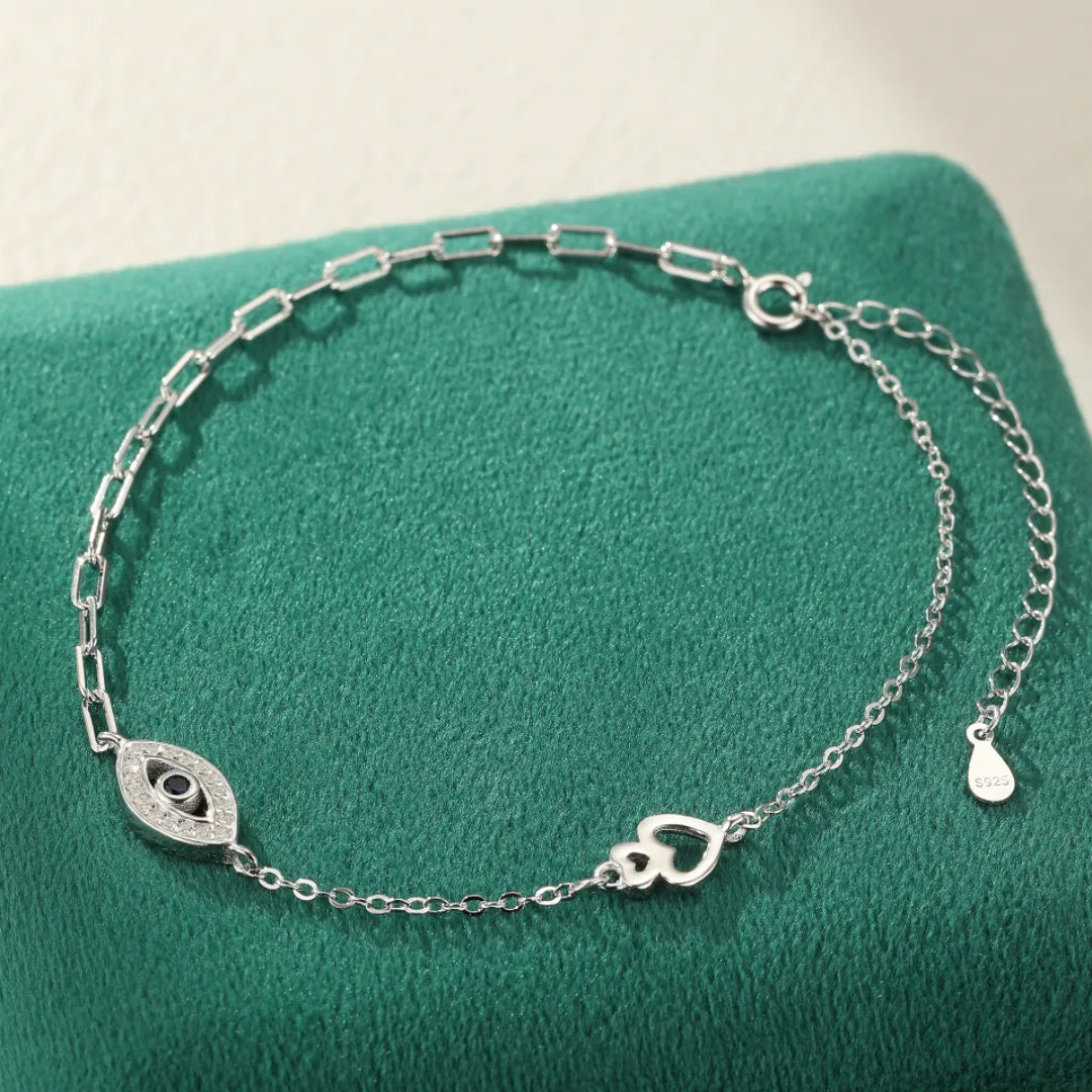Bracelet de cheville en argent 925 orné d'un œil protecteur serti d'une pierre bleue et de petits cristaux, avec un motif de deux cœurs entrelacés.