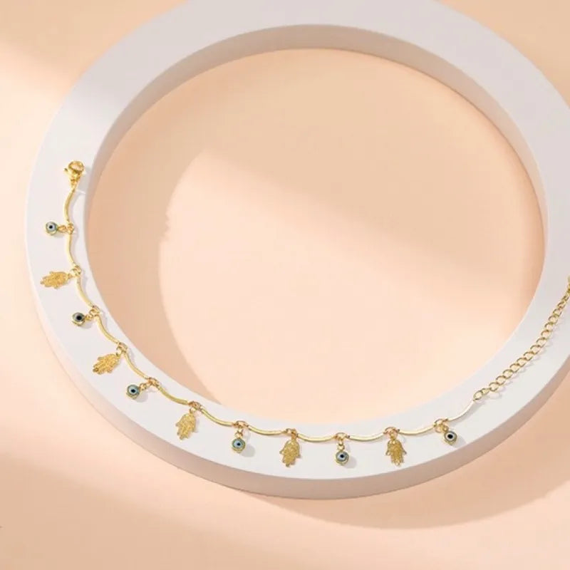 Bracelet de cheville doré avec des pendentifs en forme de main de Fatma (Khamsa) et de mauvais œil bleus alternés, sur un fond clair.