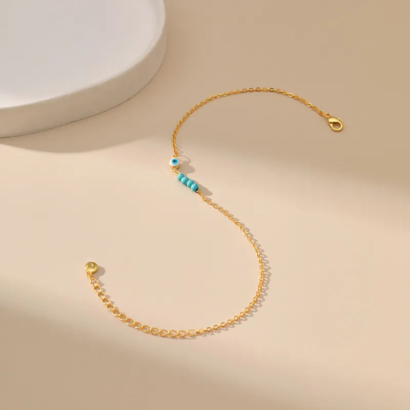 Bracelet de cheville doré avec un œil bleu et des perles bleues turquoise, symbole de protection, sur fond neutre.