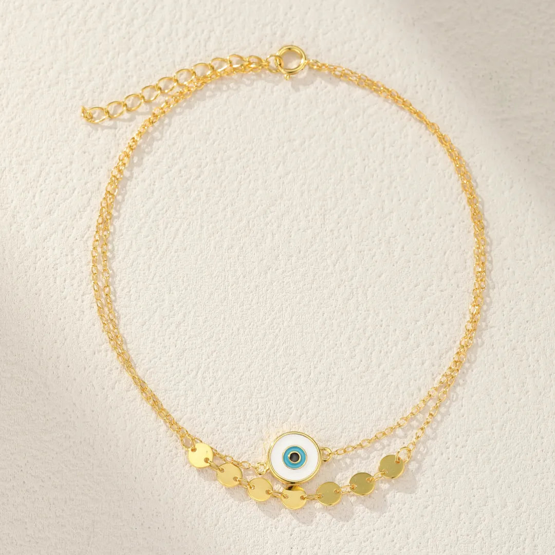 Bracelet de cheville double chaîne en argent avec un pendentif œil bleu et blanc et des breloques rondes dorées.